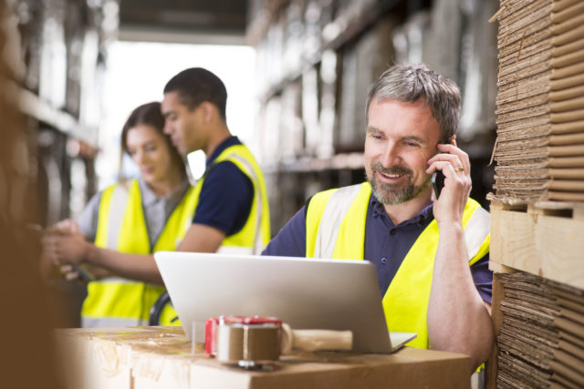 a mature warehouse manager stands in his warehouse looking at his laptop whilst talking on the telephone.He is wearing a hi vis waistcoat and stands in front of some warehouse racking. I the background two young warehouse workers can be seen .