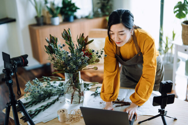 Professional young Asian female florist working on laptop while making vlog session on flower bouquet arrangement class with laptop and camera at home. Self-employment. Online classes. Small business concept